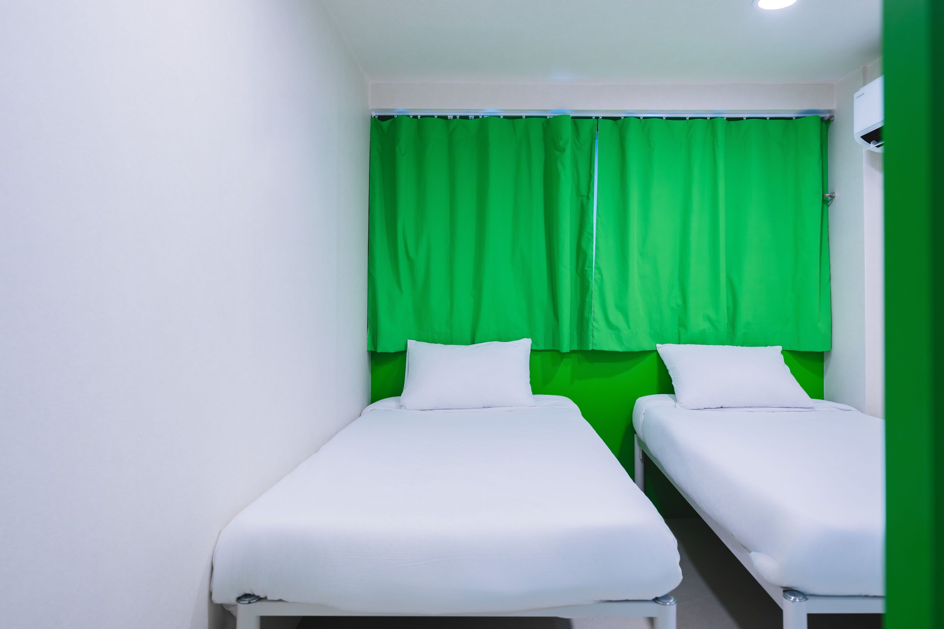 A small, narrow room with two single beds, white bedding, and green curtains.