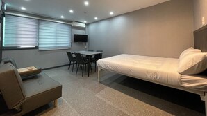 A compact hotel room with a single bed, a small desk with chairs, a television, and an air conditioning unit. - INN THE HOUR_Myeongdong (Seoul)