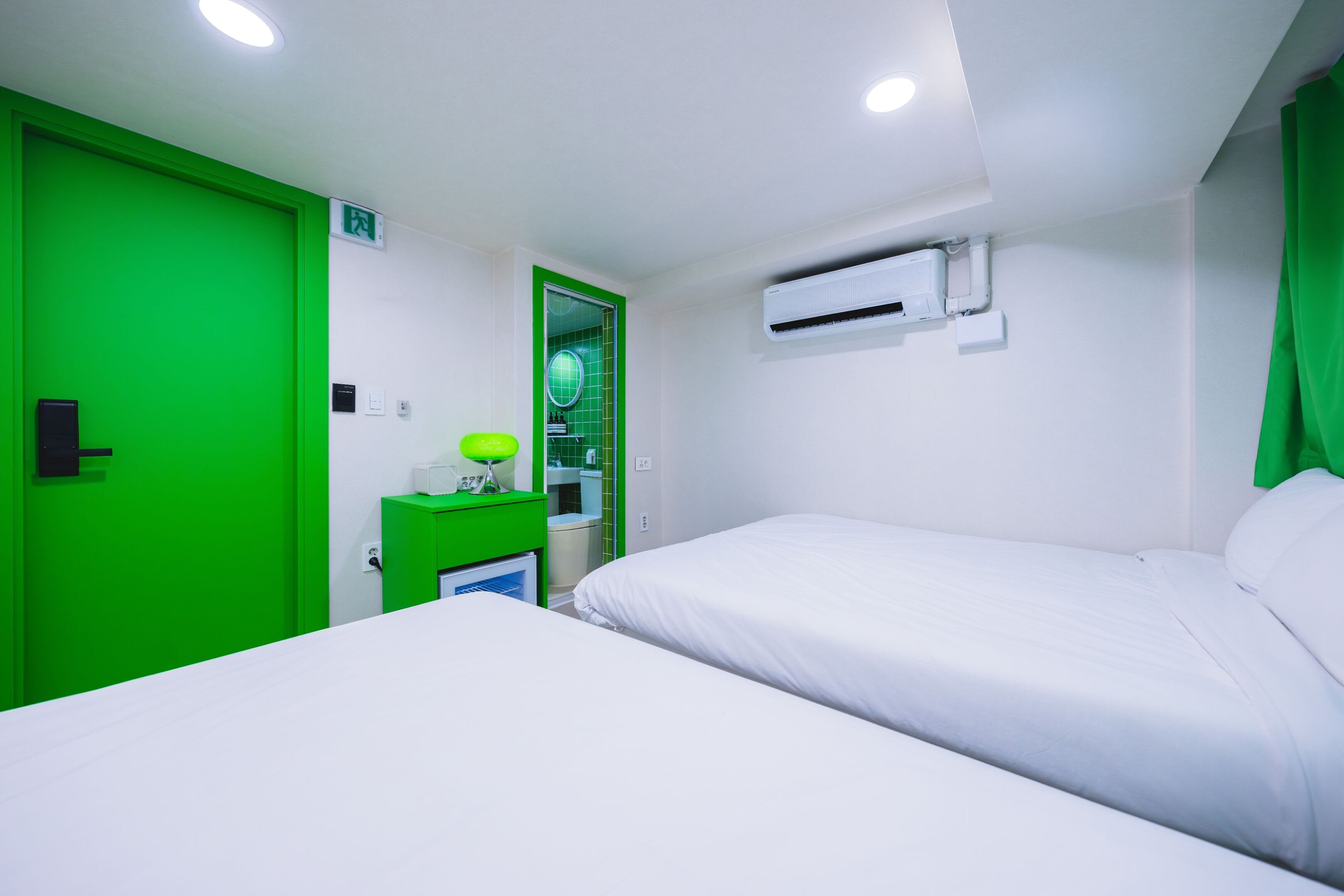 A hotel room with a bed, a green door, a small desk with a lamp, and an air conditioning unit.