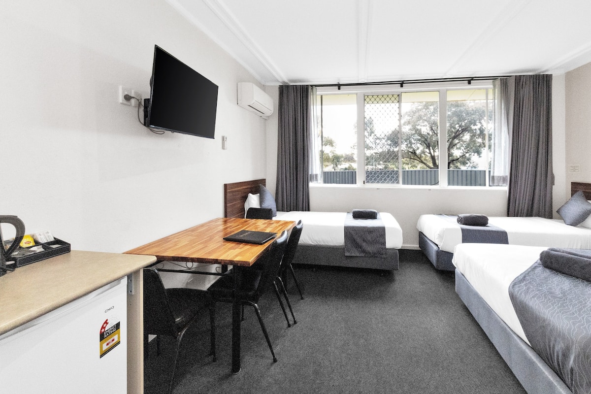 Habitación de hotel con dos camas individuales, un escritorio de madera con silla, un televisor de pantalla plana montado en la pared y una ventana con cortinas.