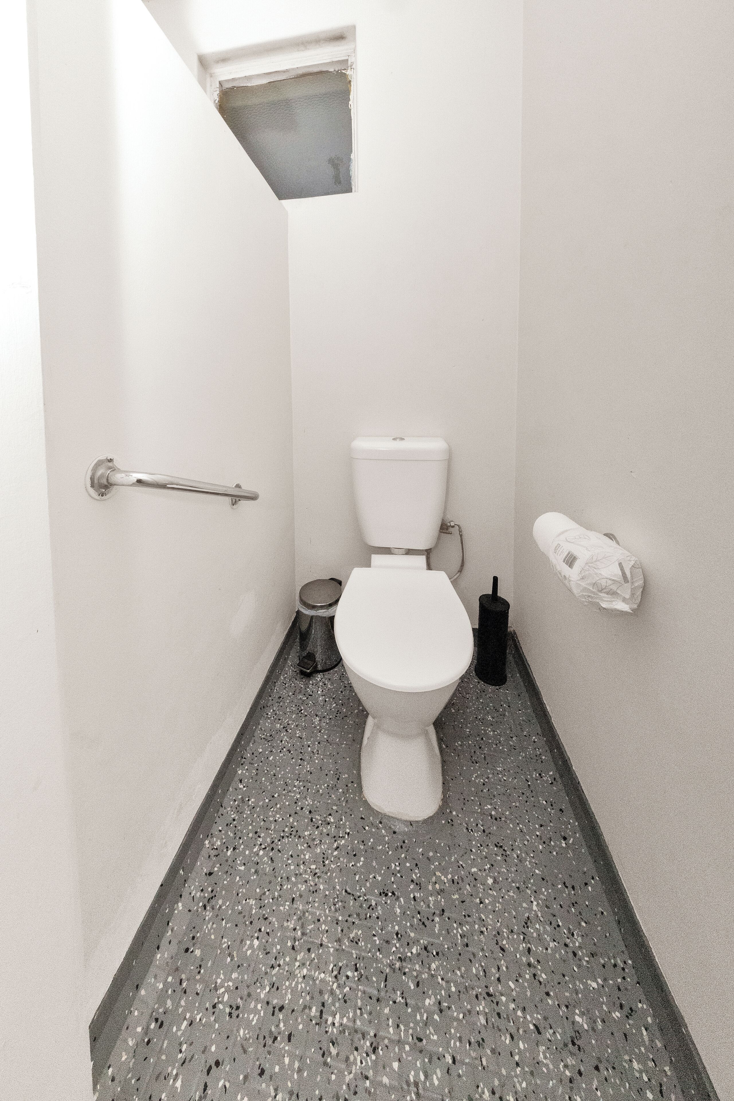A small, narrow bathroom with a white toilet, a handrail, a trash bin, and a paper towel dispenser on the wall.