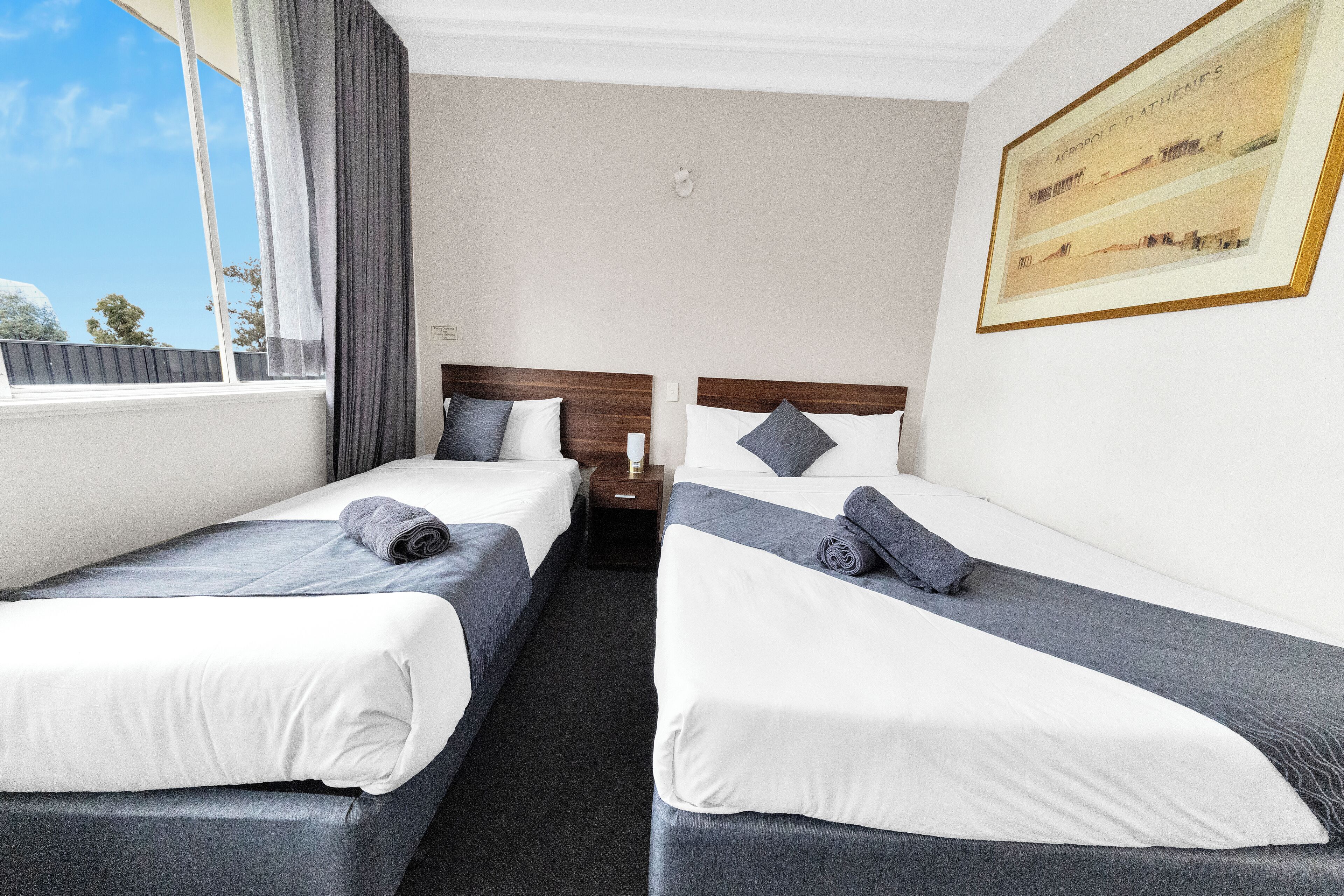 Quarto de hotel com duas camas, uma janela grande com cortinas, um quadro emoldurado na parede e uma mesa de cabeceira com abajur.