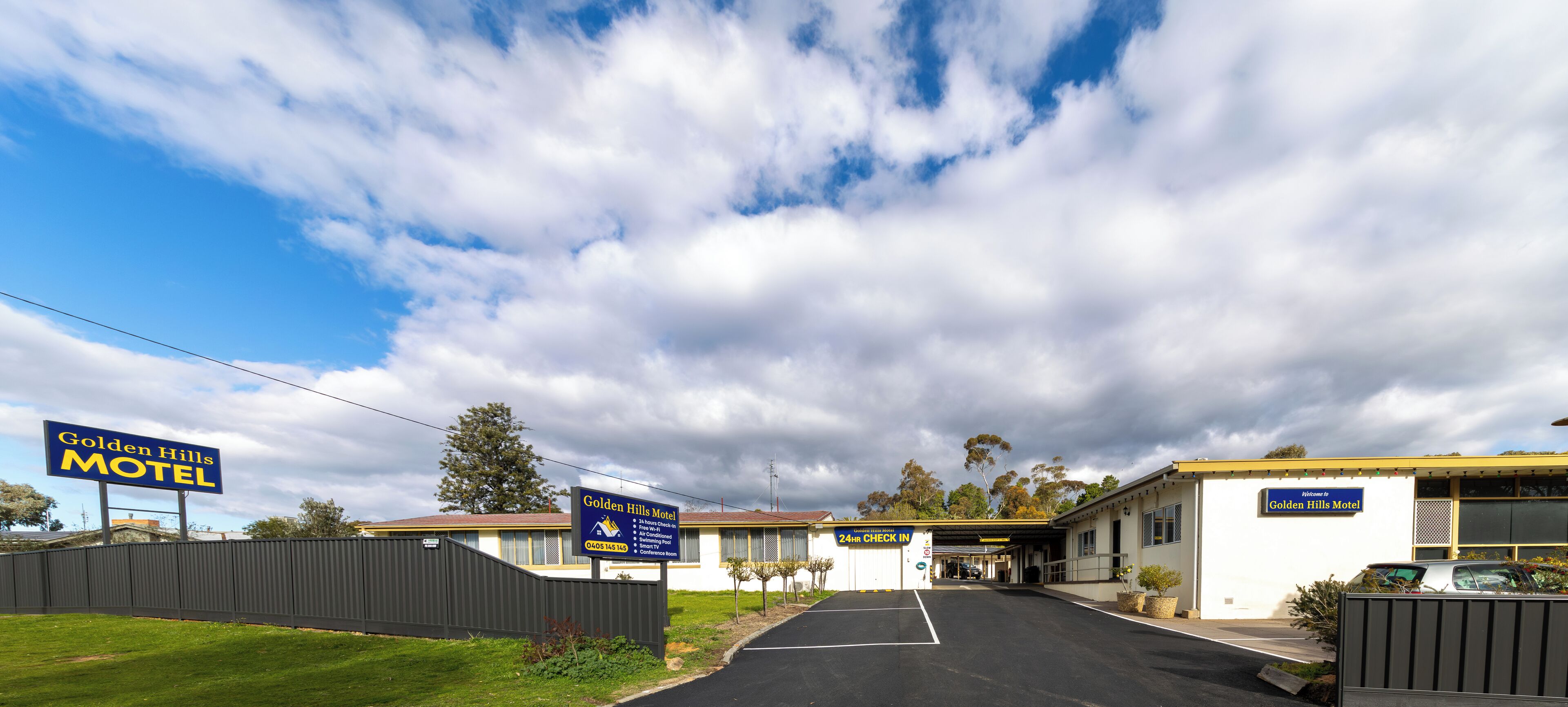 A motel named Golden Hills Motel with a 24-hour check-in, a fenced parking area, and a clear sky with clouds.