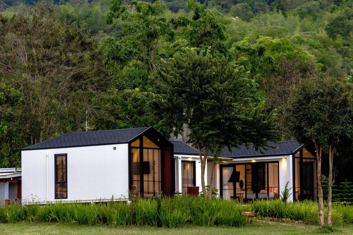 Een modern, twee verdiepingen tellend huis met grote ramen en een plat dak, omgeven door weelderig groen en bomen.