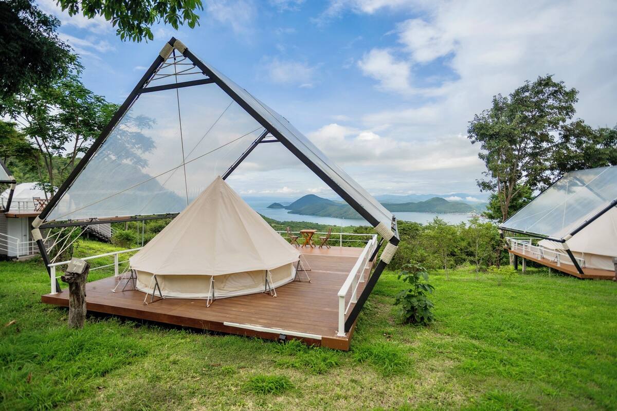 Een geodetische koepeltent met een houten terras en een tafel, voor een achtergrond van bergen en een meer.