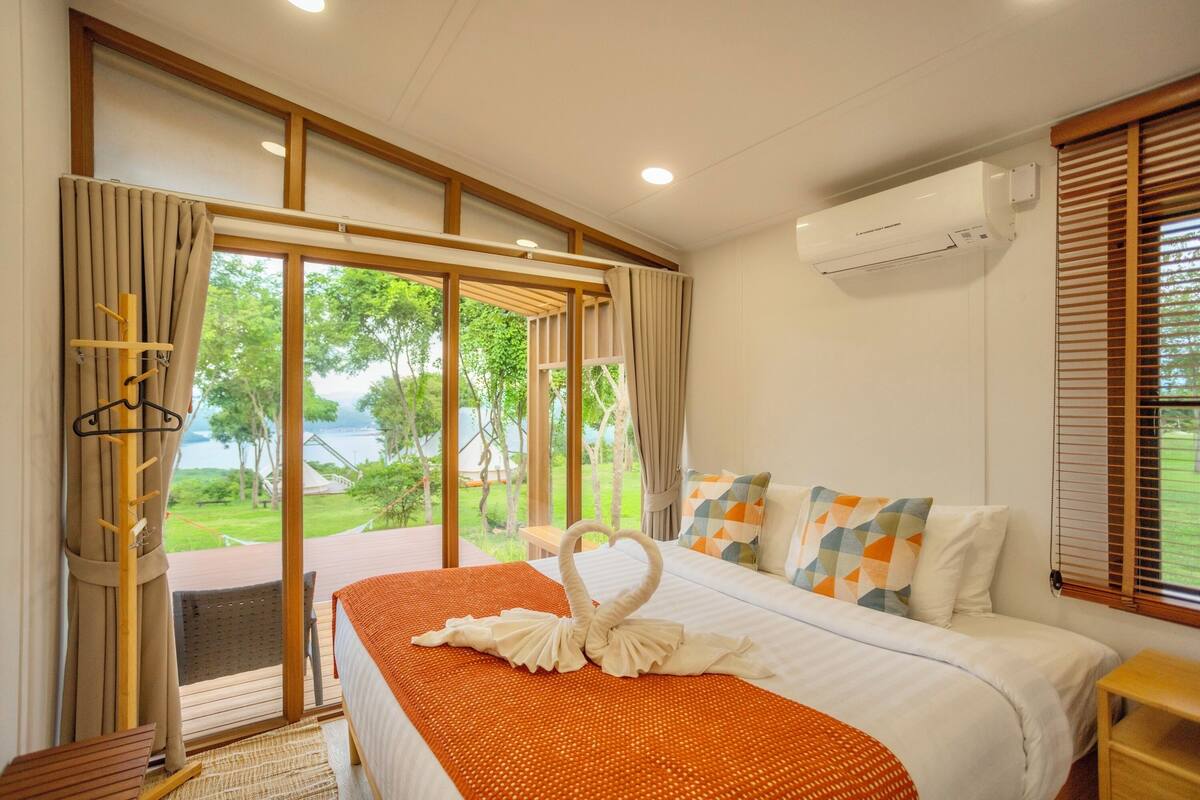 Hotelkamer met een bed, een raam met jaloezieën, een wandairconditioner en uitzicht op groen.