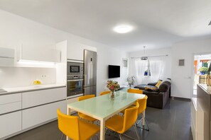 A modern kitchen with white cabinets, a stainless steel refrigerator, and a dining area with a glass table and yellow chairs.