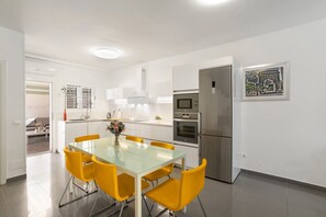 A modern kitchen with stainless steel appliances, a glass dining table with yellow chairs, and a framed picture on the wall.
