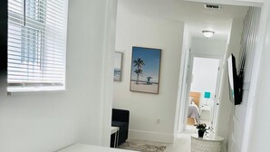 A modern, minimalist room with a white desk, a small table, and a chair. There is a painting on the wall and a potted plant on a round table.