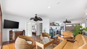 Un salon spacieux avec un grand canapé, une table basse en verre et des fauteuils en osier. Un téléviseur à écran plat est fixé au mur, et un ventilateur de plafond est installé au plafond.