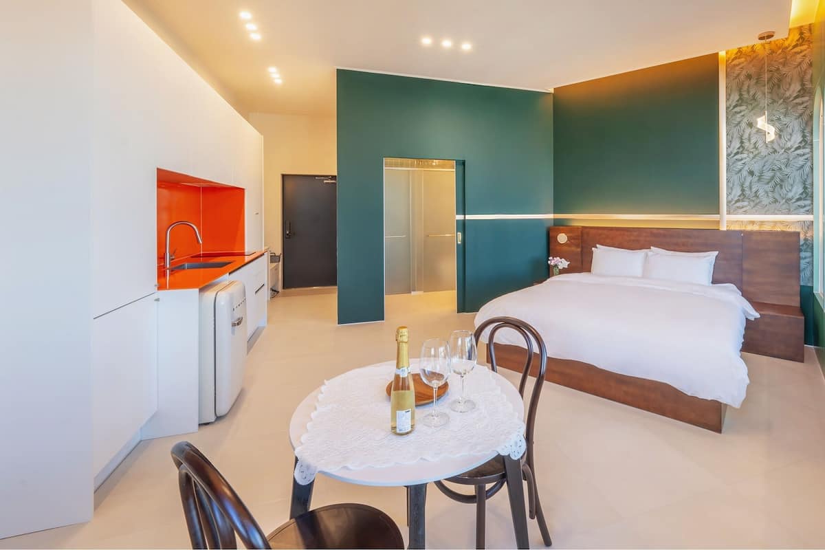 A modern hotel room with a bed, a small kitchenette, a dining table with chairs, and a glass-enclosed bathroom.