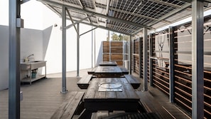 Un patio al aire libre con mesas y bancos de madera, una pérgola de metal y un área de almacenamiento con estantes.