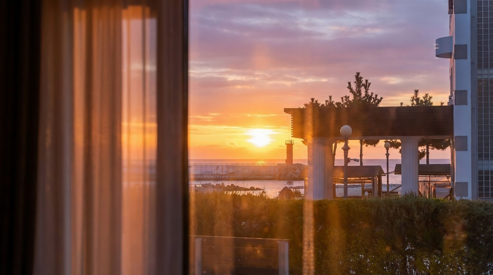 A sunset view from a window overlooking a coastal area with a gazebo and a building.
