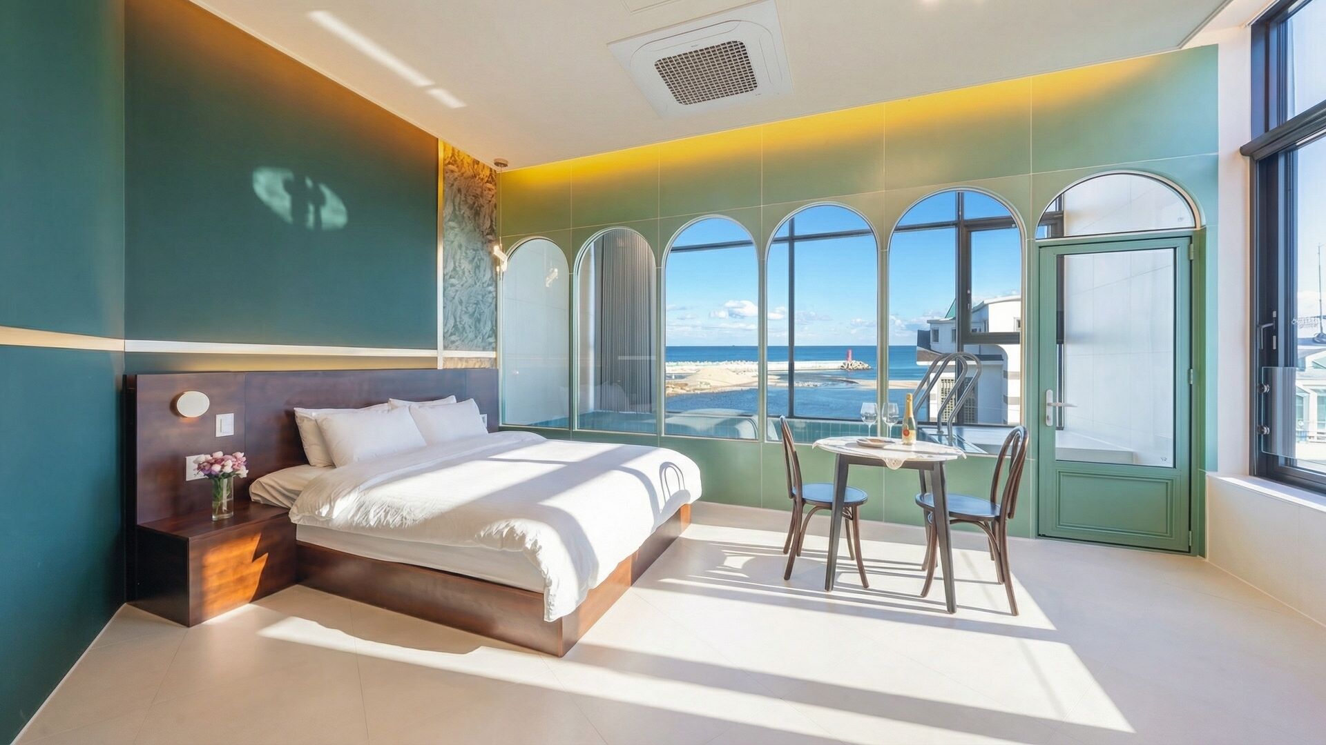 A modern bedroom with a bed, a small table with chairs, and a view of the beach through large windows.