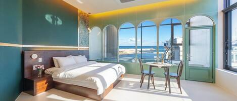 A modern bedroom with a bed, a small table with chairs, and a view of the beach through large windows.