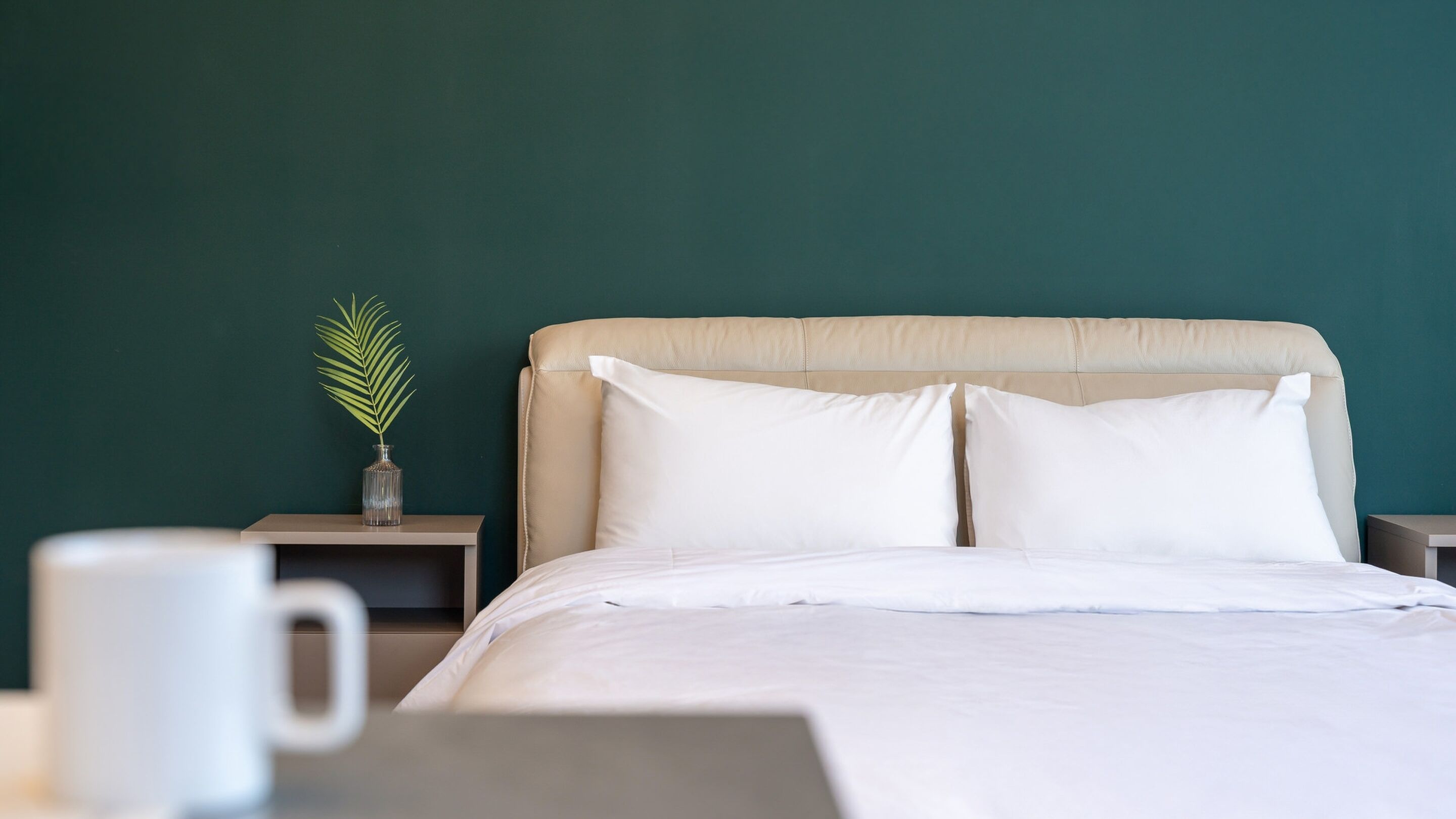 A neatly made bed with white linens, a beige headboard, and two bedside tables with a small vase of green foliage.
