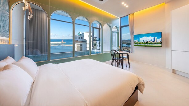 Ein modernes Hotelzimmer mit einem großen Bett, einem Essbereich mit Tisch und Stühlen sowie großen Fenstern mit Blick auf das Meer und Gebäude.