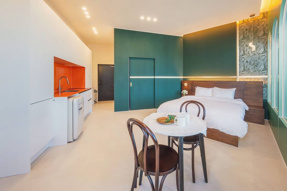 A compact hotel room with a kitchenette, a bed with white linens, a small round table with two chairs, and a window with a view of the city.