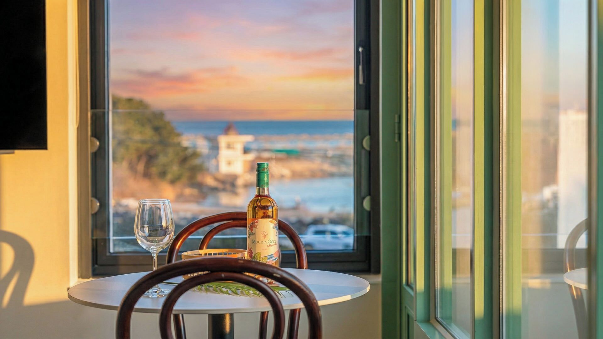 A table with a bottle of wine and a glass, positioned by a window overlooking a coastal view.