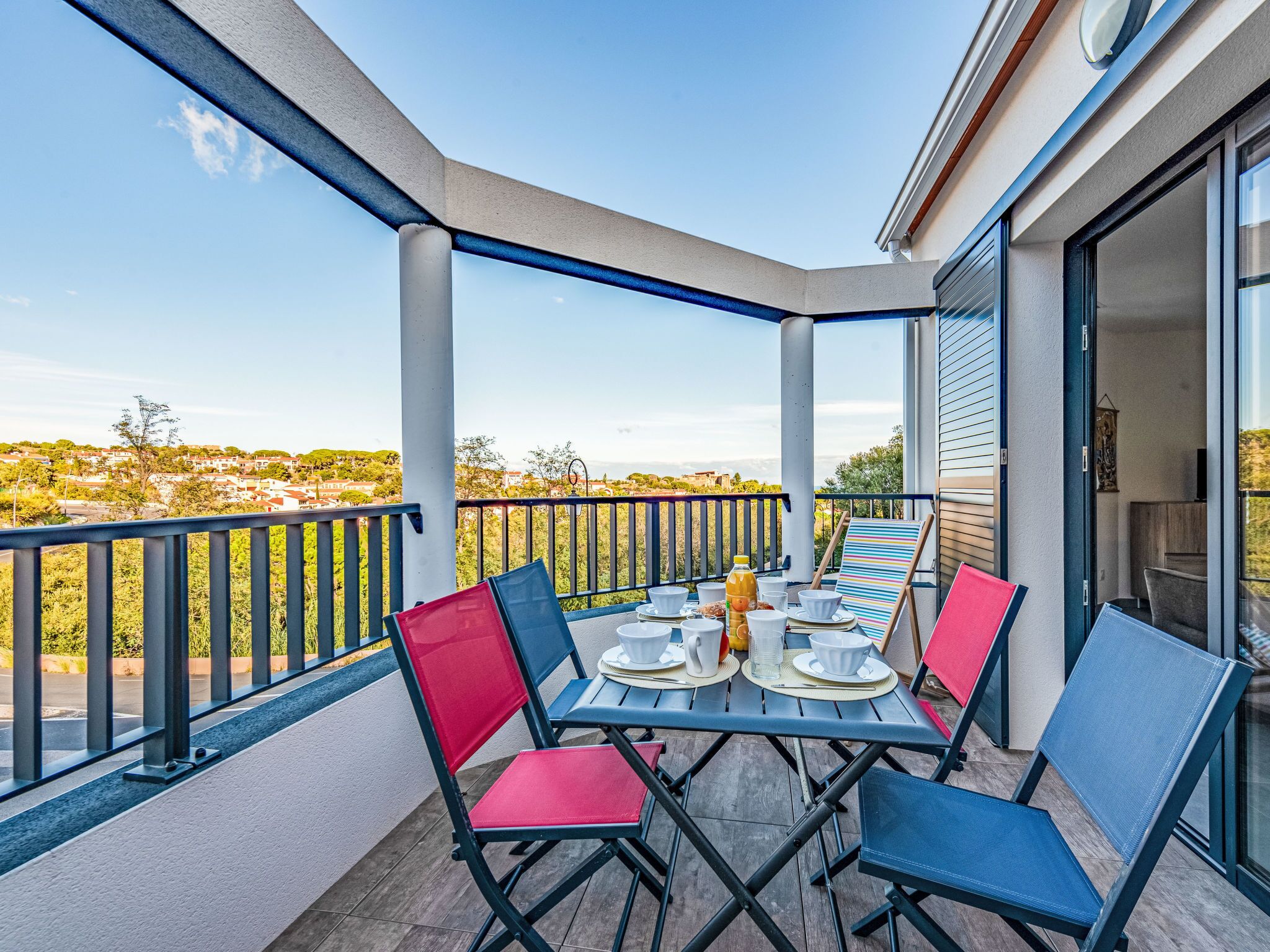 Un espacio cubierto para comer al aire libre con una mesa dispuesta para cuatro personas, sillas alrededor y vista a una zona residencial al fondo.