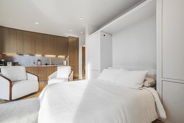 A modern bedroom with a large bed, two white armchairs, a kitchen area with wooden cabinets, and a white wall. - Luxury new studio in downtown Healdsburg with WiFi, AC, Poolhouse/Fitness Center (Healdsburg)