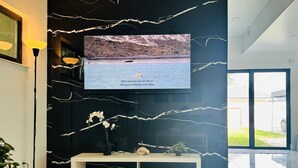 Moderni olohuone, jossa on suuri, seinälle asennettu litteänäyttöinen televisio. Televisio on kiinnitetty mustaan seinään, jossa on valkoisia, halkeamia muistuttavia kuvioita. Etualalla on valkoinen konsolipöytä, jonka päällä on koriste-esineitä ja kulho.