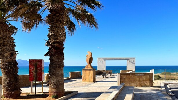 A coastal promenade with palm trees, a sculpture, and a view of the sea.