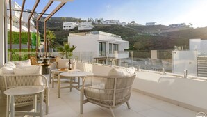 A balcony with wicker furniture, a glass table, and a view of modern buildings on a hillside. - Villa Kolibri | Mountain view - pool - garden (La Cala de Mijas)