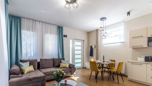 A modern living room with a sofa, dining table, and chairs, a kitchen area, and a large window with curtains.