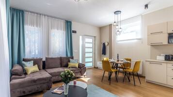 A modern living room with a sofa, dining table, and chairs, a kitchen area, and a large window with curtains.