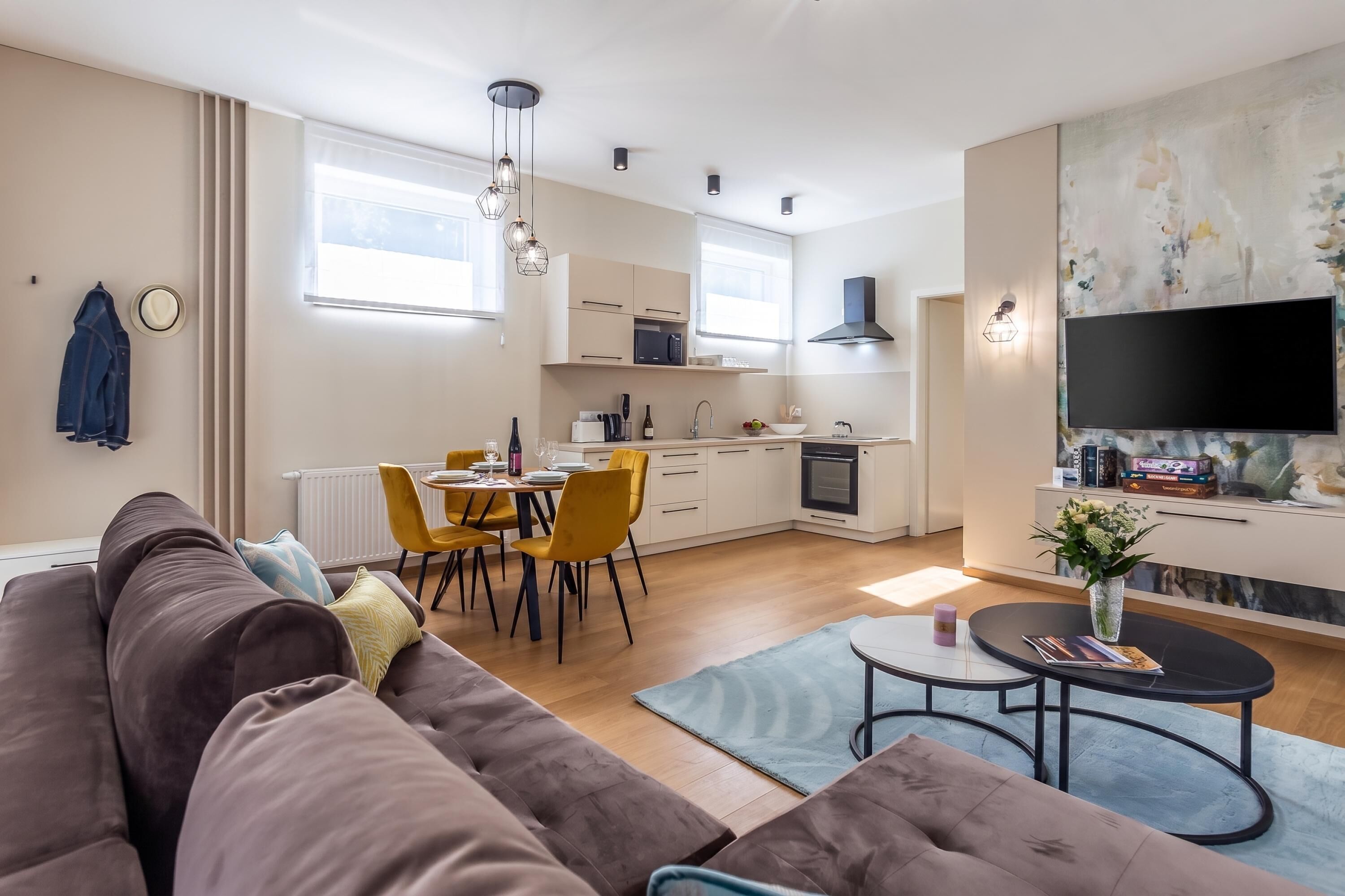 A modern living room with a sofa, dining table, and chairs, a kitchen with appliances, and a TV mounted on the wall.