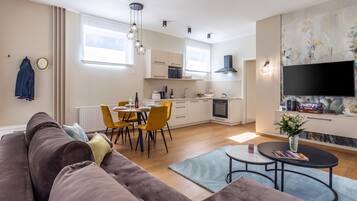 A modern living room with a sofa, dining table, and chairs, a kitchen with appliances, and a TV mounted on the wall.