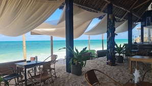 A beachside restaurant with outdoor seating, a thatched roof, and a view of the ocean. - Garden Beach Bungalows (Jambiani)