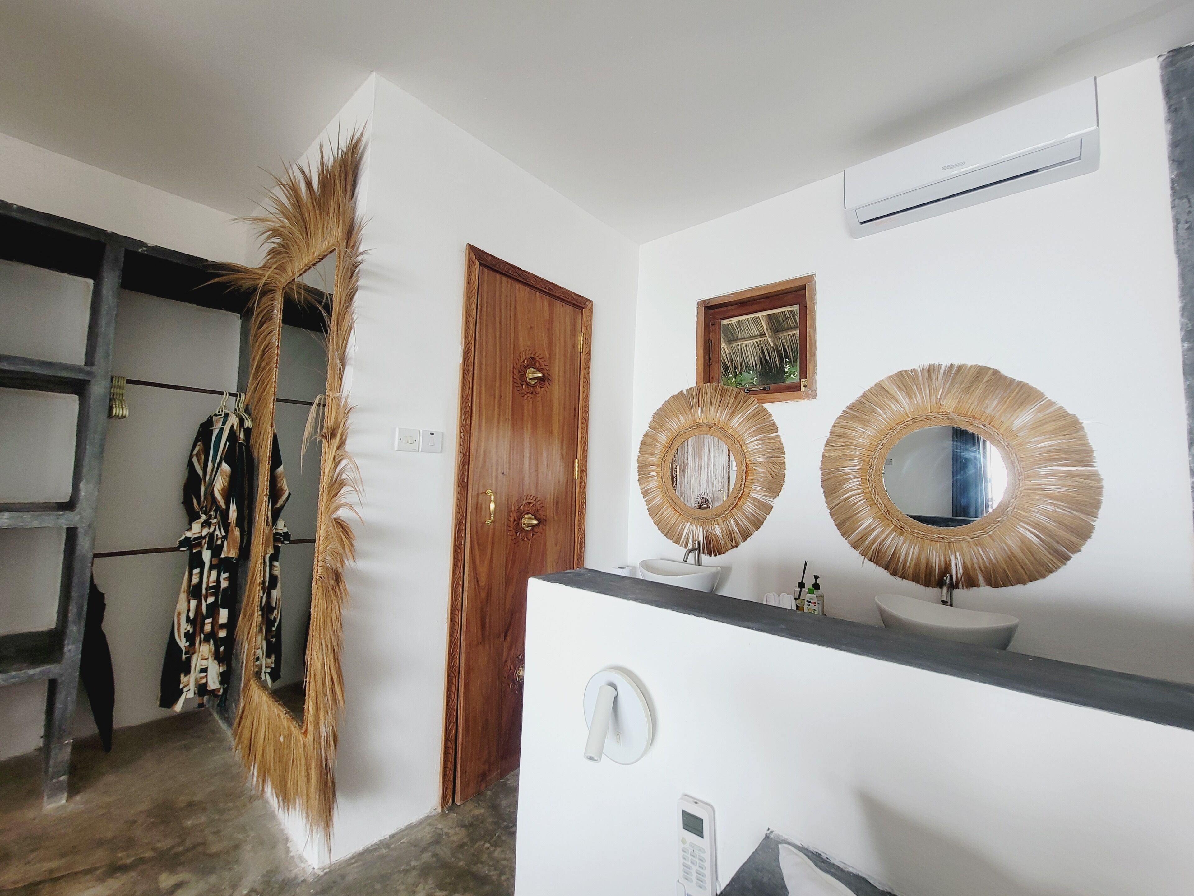 A bathroom with a wooden door, a white sink, two round mirrors with straw-like frames, and a small window with a thatched roof view.