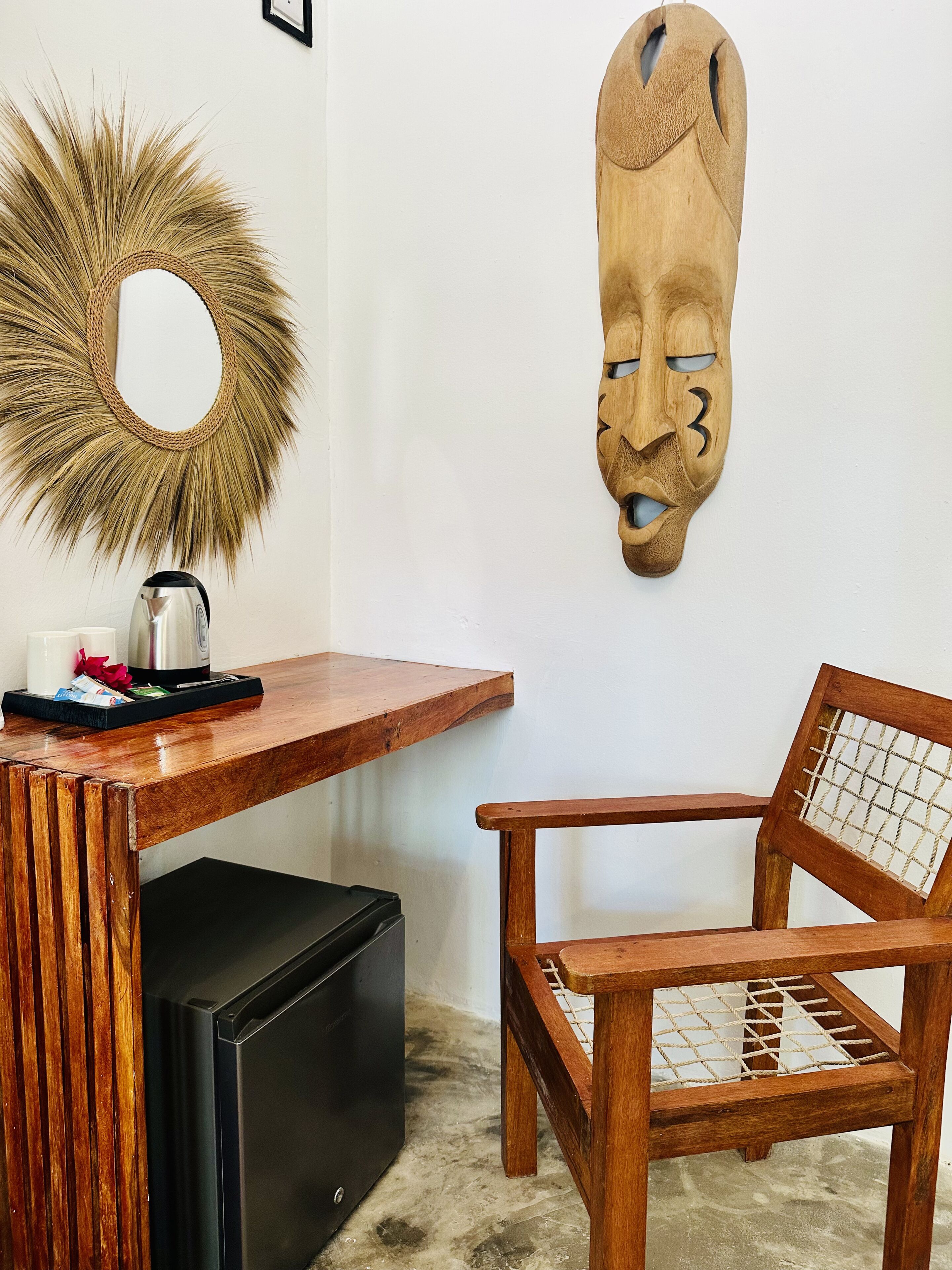 A wooden desk with a woven mirror, a chair with a woven backrest, and a black refrigerator.