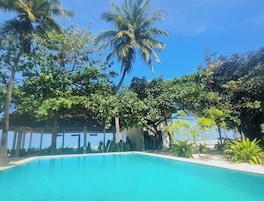 A clear blue pool surrounded by lush greenery and palm trees, with a covered seating area in the background.