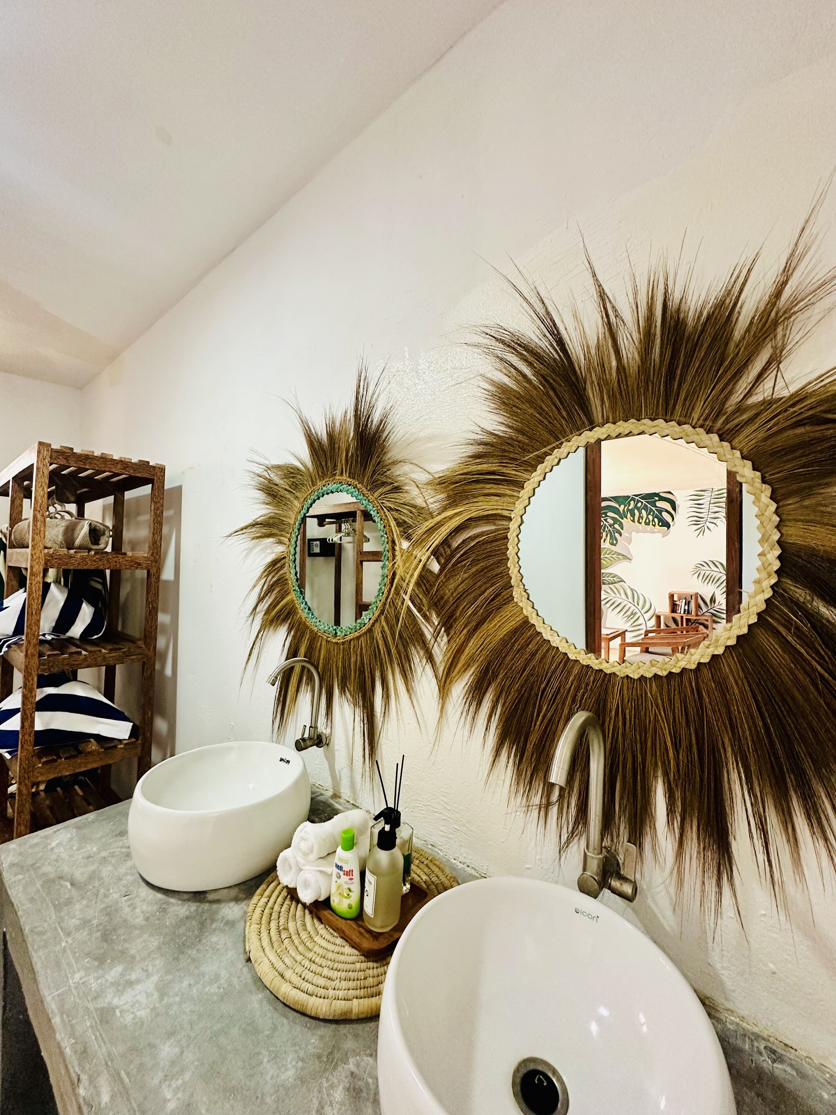 Un baño con dos espejos, un estante de madera con toallas y un lavamanos con artículos de tocador.