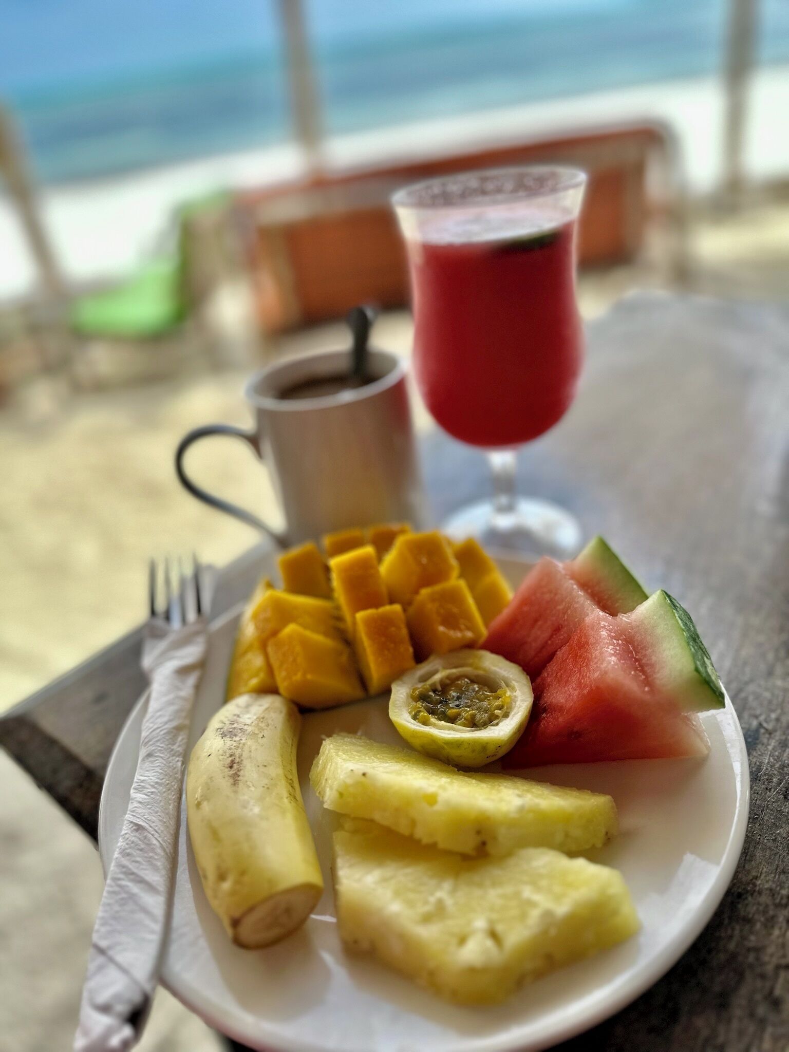 Un plat amb fruita i una beguda sobre una taula al costat d'una finestra que dona a la platja.