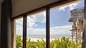 A window with a view of a beach, a thatched structure, and green foliage. - Garden Beach Bungalows (Jambiani)