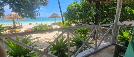 A beach view from a covered area with a wooden railing, palm trees, and a thatched roof structure.