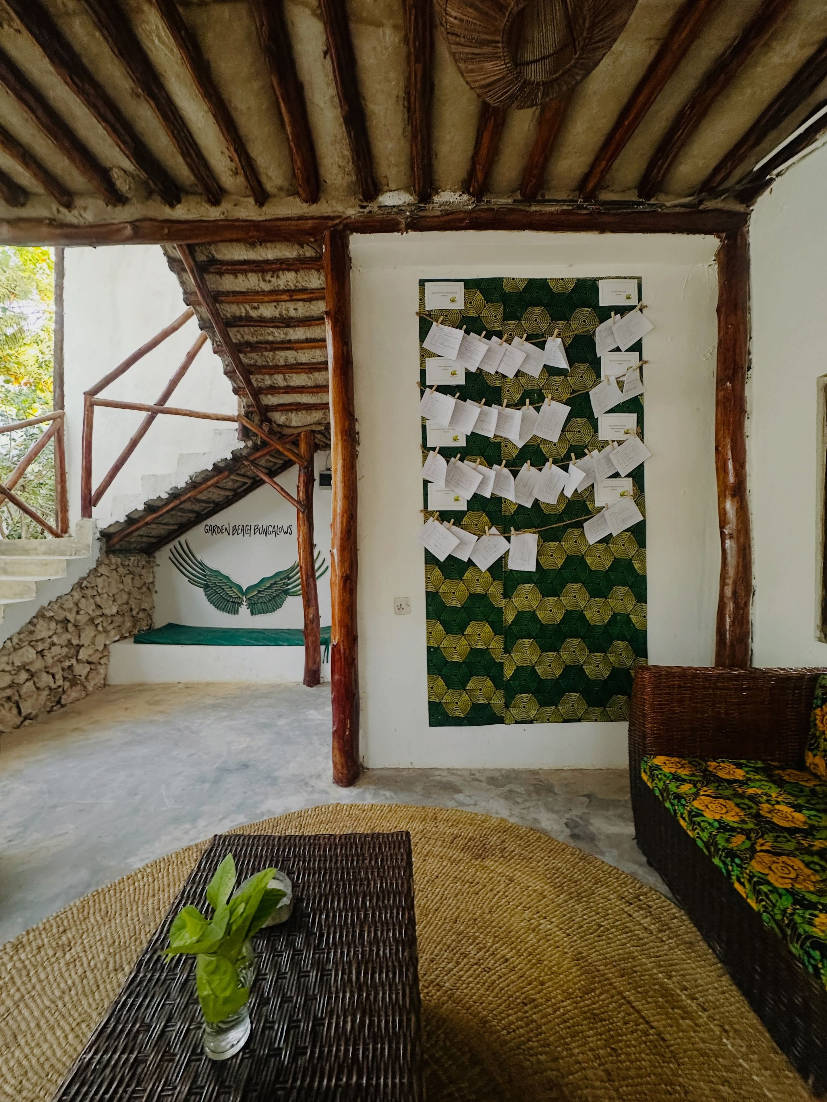 A room with a staircase, a wall with pinned papers, a woven mat, and a wicker sofa.