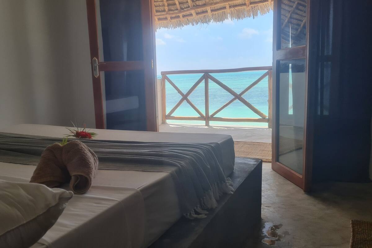 Una habitació amb un llit, vista al mar a través d'una porta oberta i una estructura de teules.