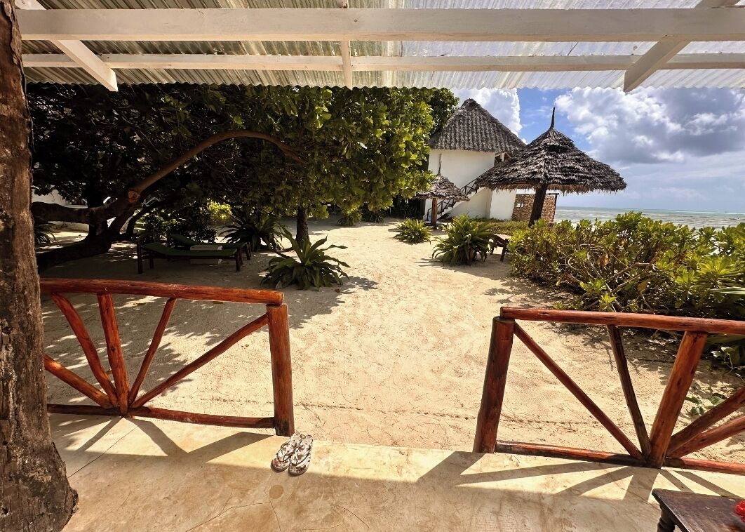 Una propiedad frente al mar con un portón de madera, una estructura de techo de paja y un sendero arenoso que conduce a un edificio.