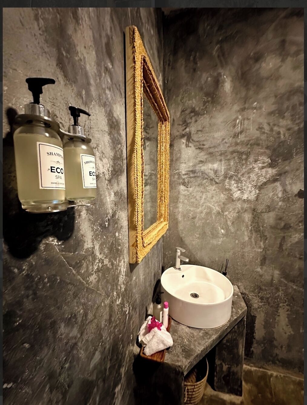 A bathroom with a stone wall, a wooden shelf holding a white sink, a mirror, and two bottles of shampoo.