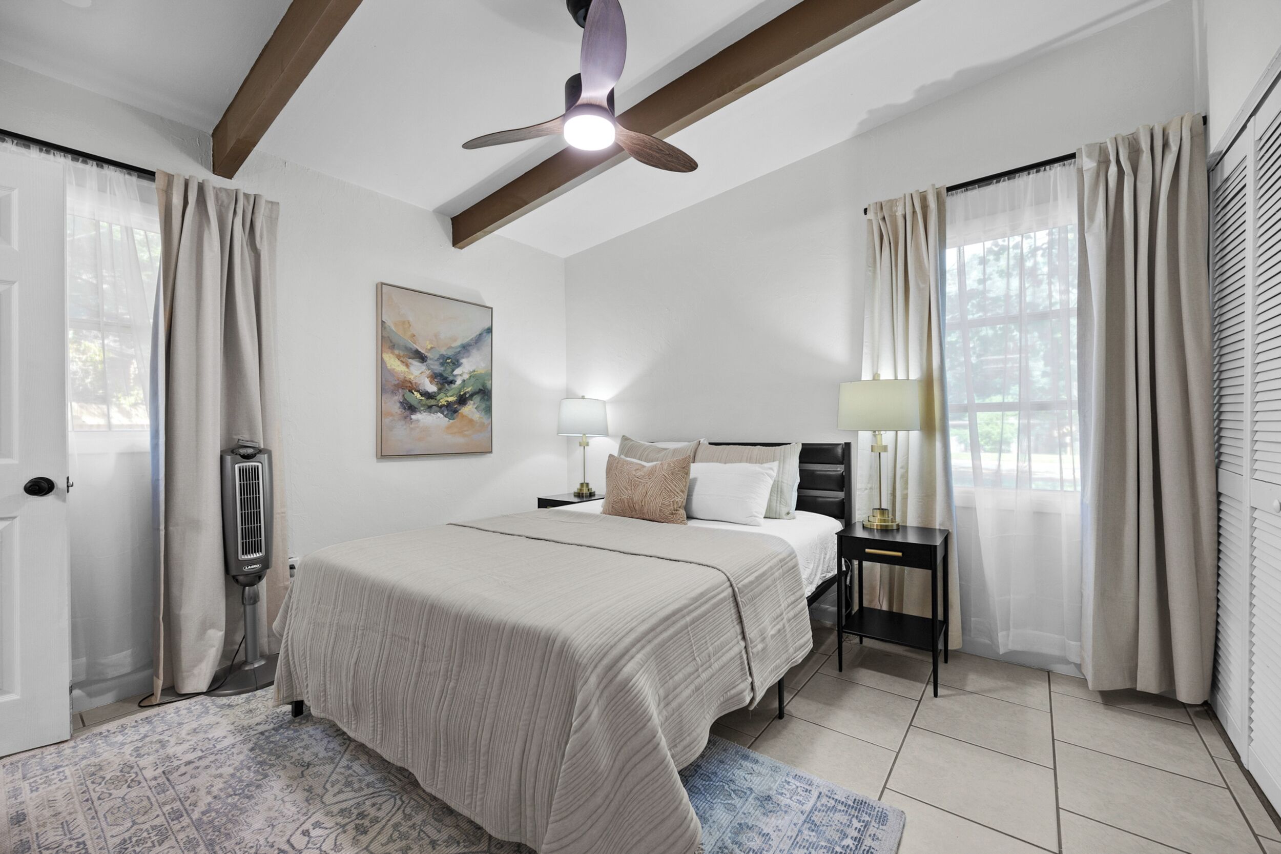 Quarto com cama, mesas de cabeceira, ventoinha no teto, janela com cortinas e radiador.