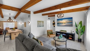 Ein modernes Wohnzimmer mit grauem Sofa, Holzbalken, einem Essbereich mit Tisch und Stühlen und einem großen Gemälde eines Sonnenuntergangs.