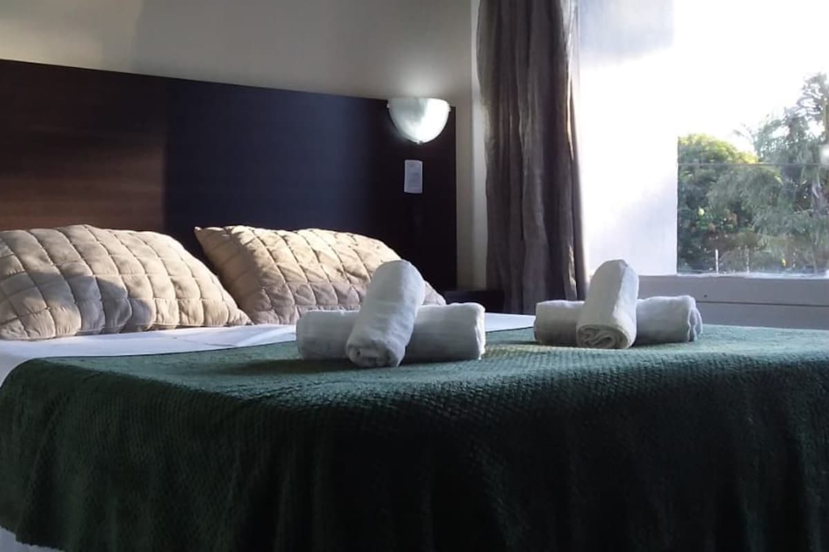 Una habitación de hotel con una cama, almohadas, una manta verde y toallas.