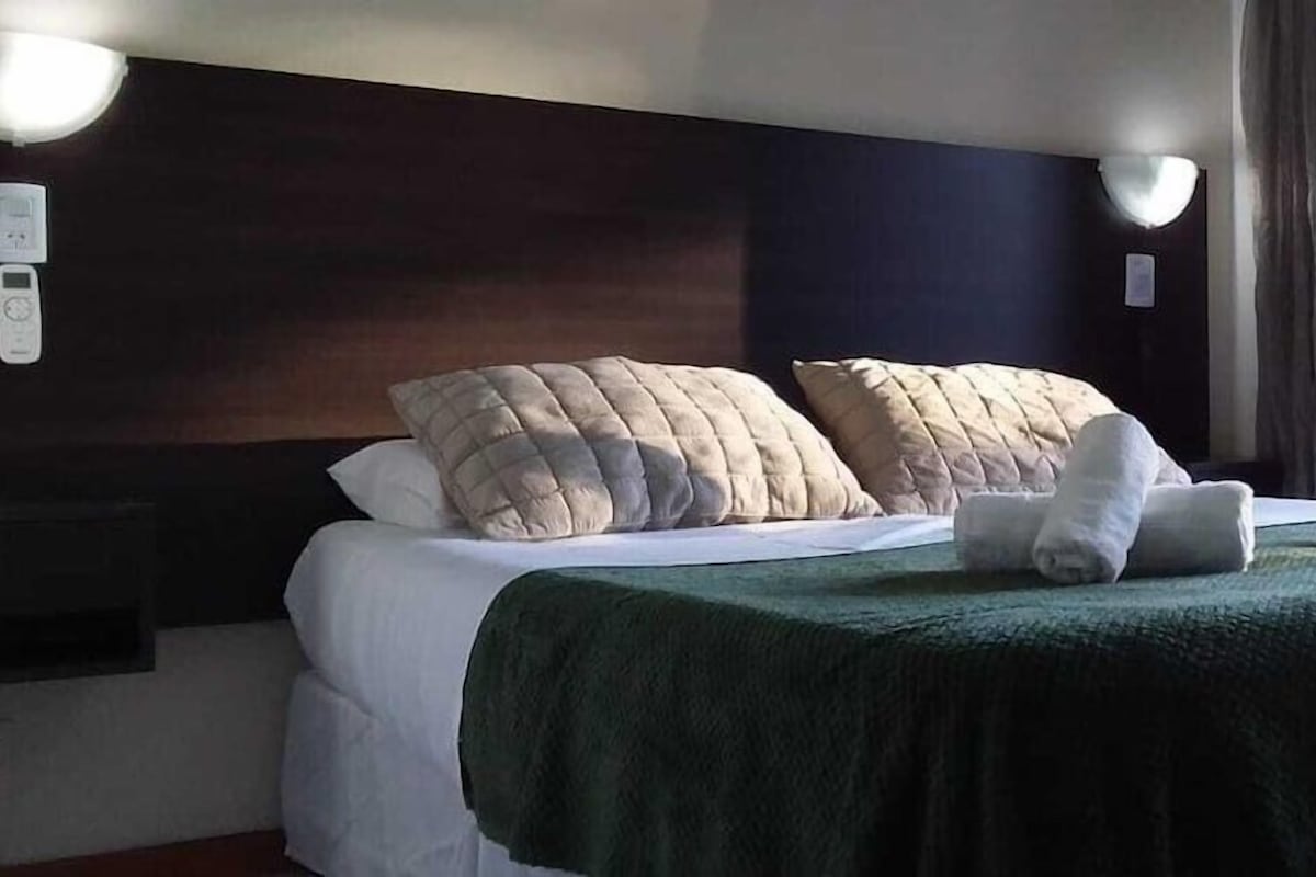 A hotel room with a bed, pillows, a green bedspread, and a window with curtains.