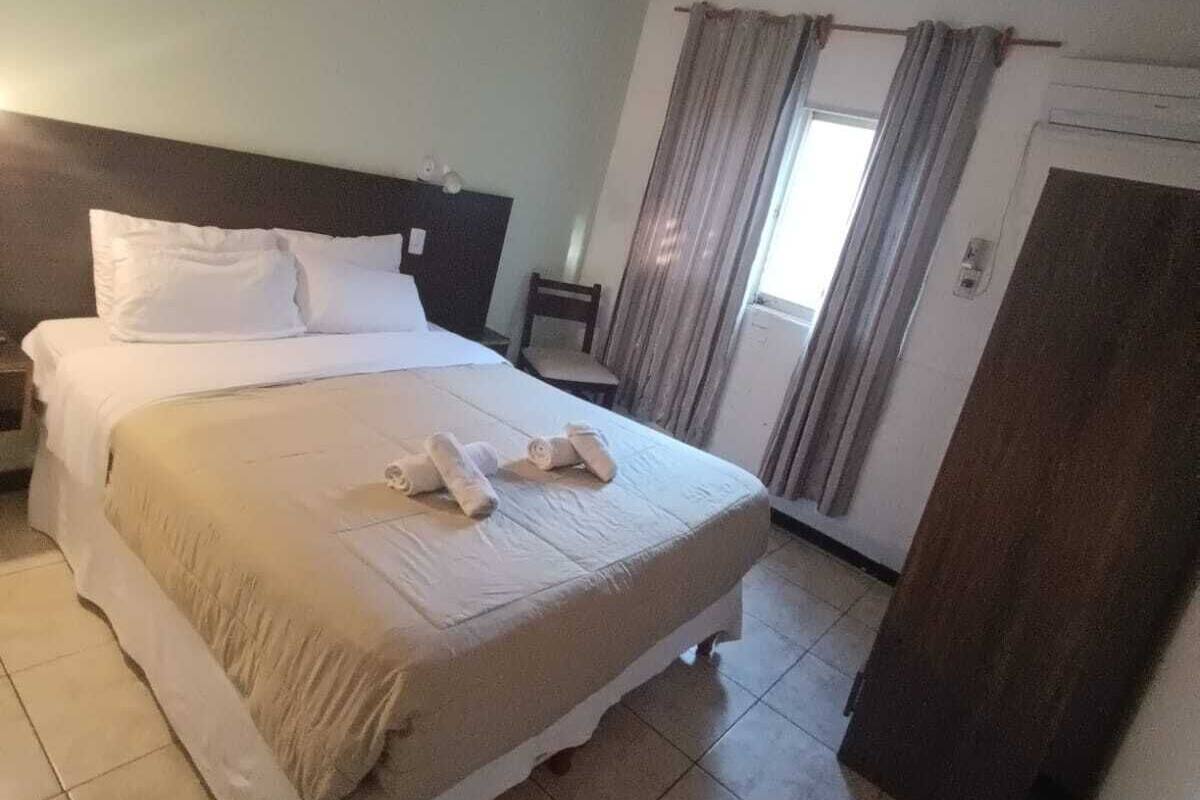 A hotel room with a bed, a nightstand, a chair, a window with curtains, and a door.