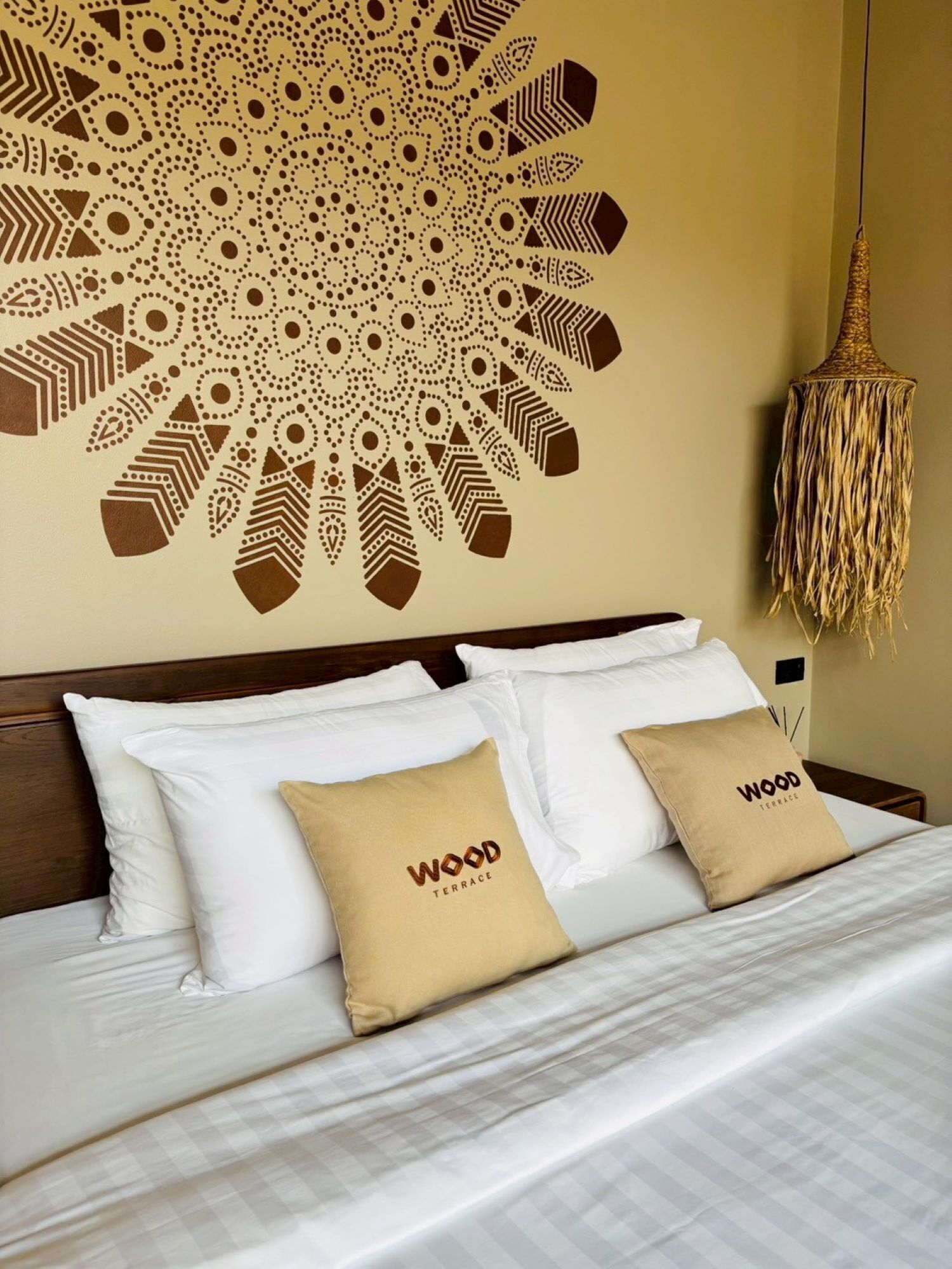 A neatly made bed with white and beige pillows, a decorative wall mural, and a woven hanging light fixture.